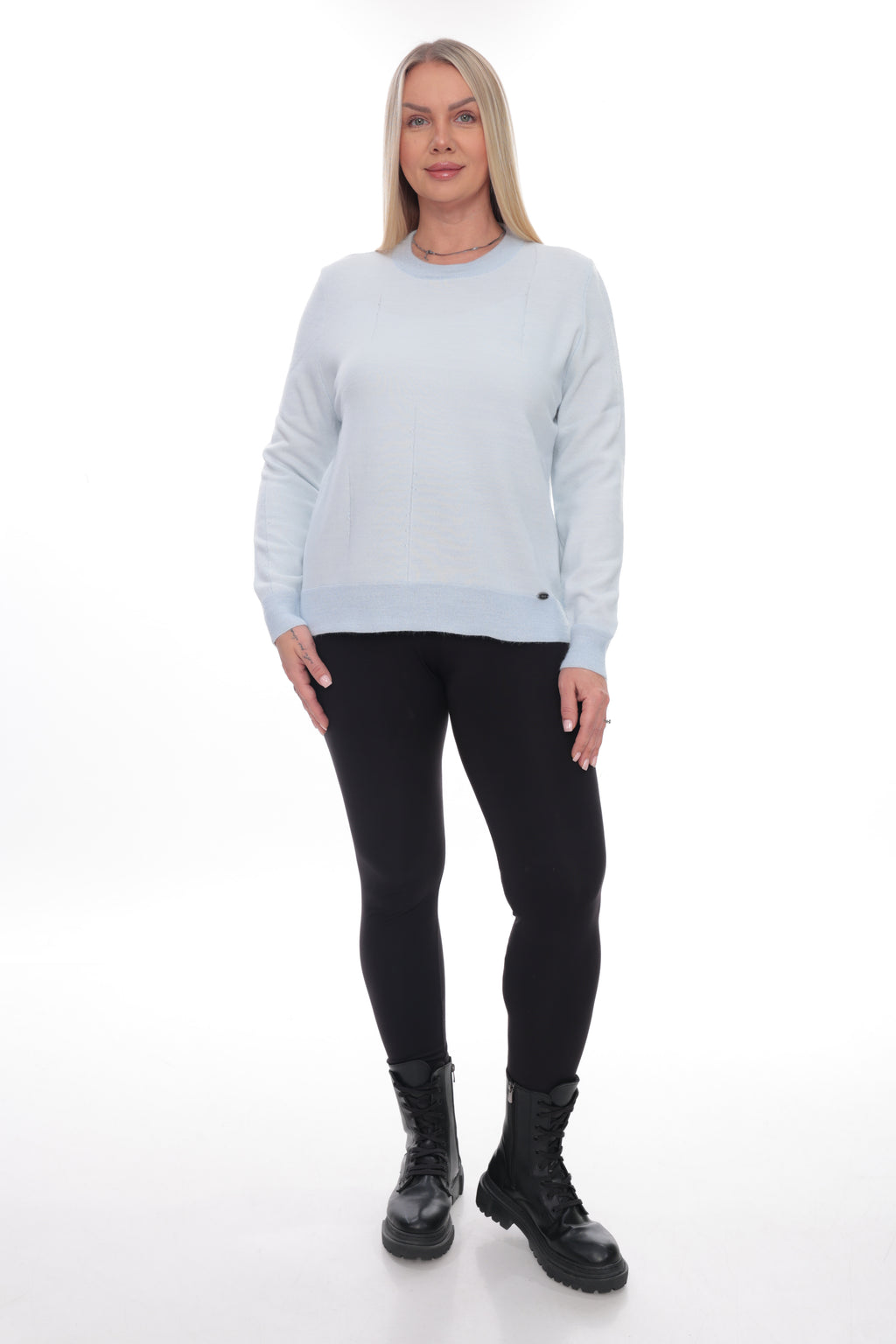A woman modeling a light blue crewneck sweatshirt with black leggings and black combat boots, standing against a white studio background.