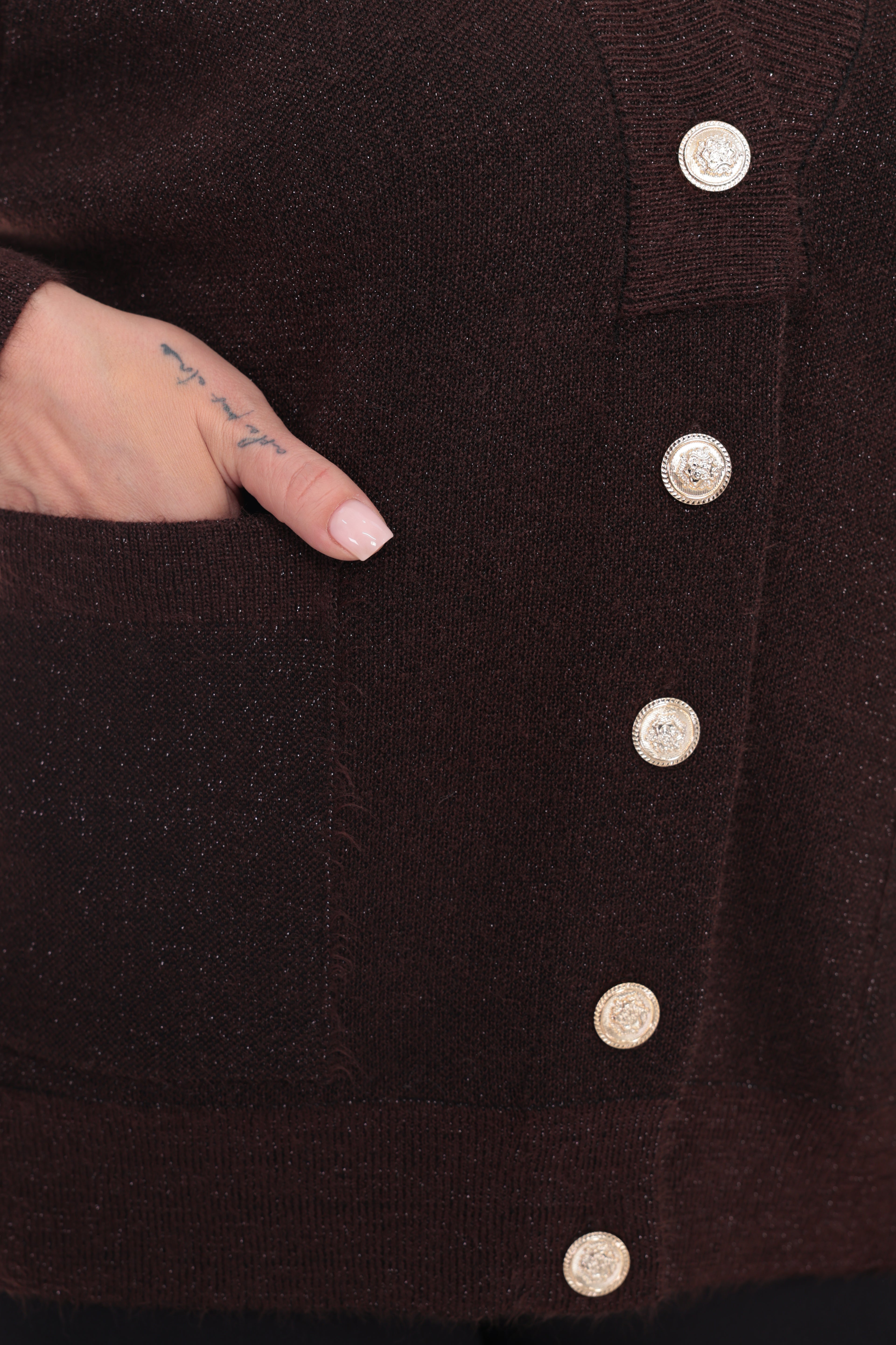 Close-up of a dark brown knit cardigan with a front pocket and decorative round metallic buttons, shown with a hand reaching into the pocket.