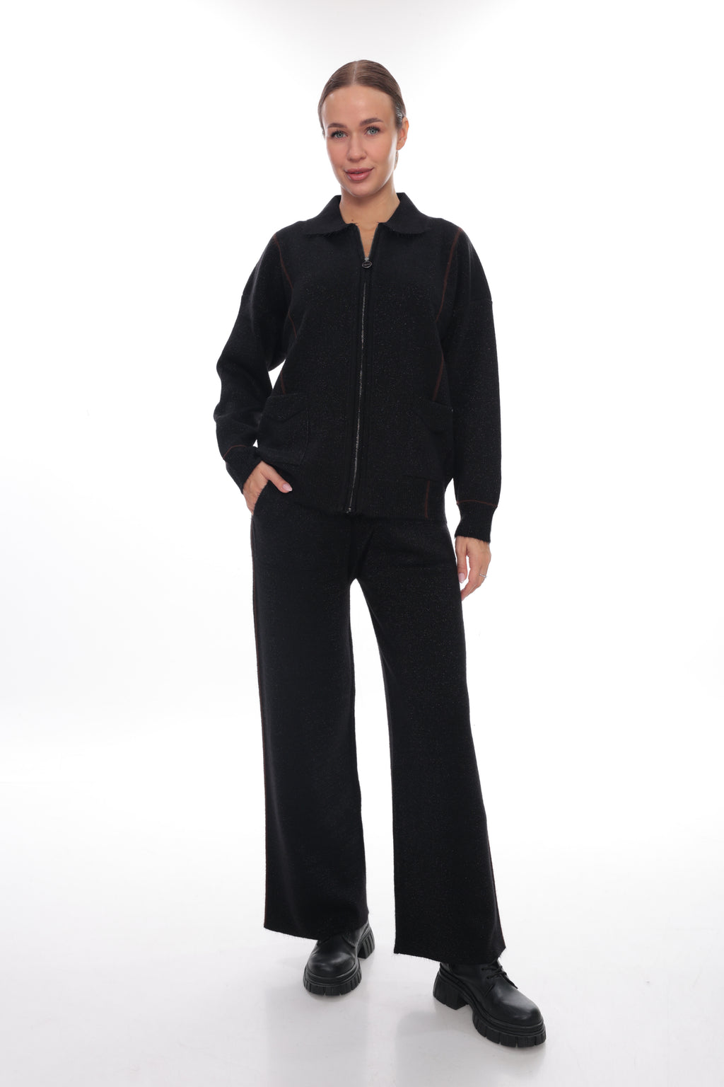 A woman modeling a black zip-up jacket and matching wide-leg trousers set, paired with black leather shoes, against a seamless white studio background.