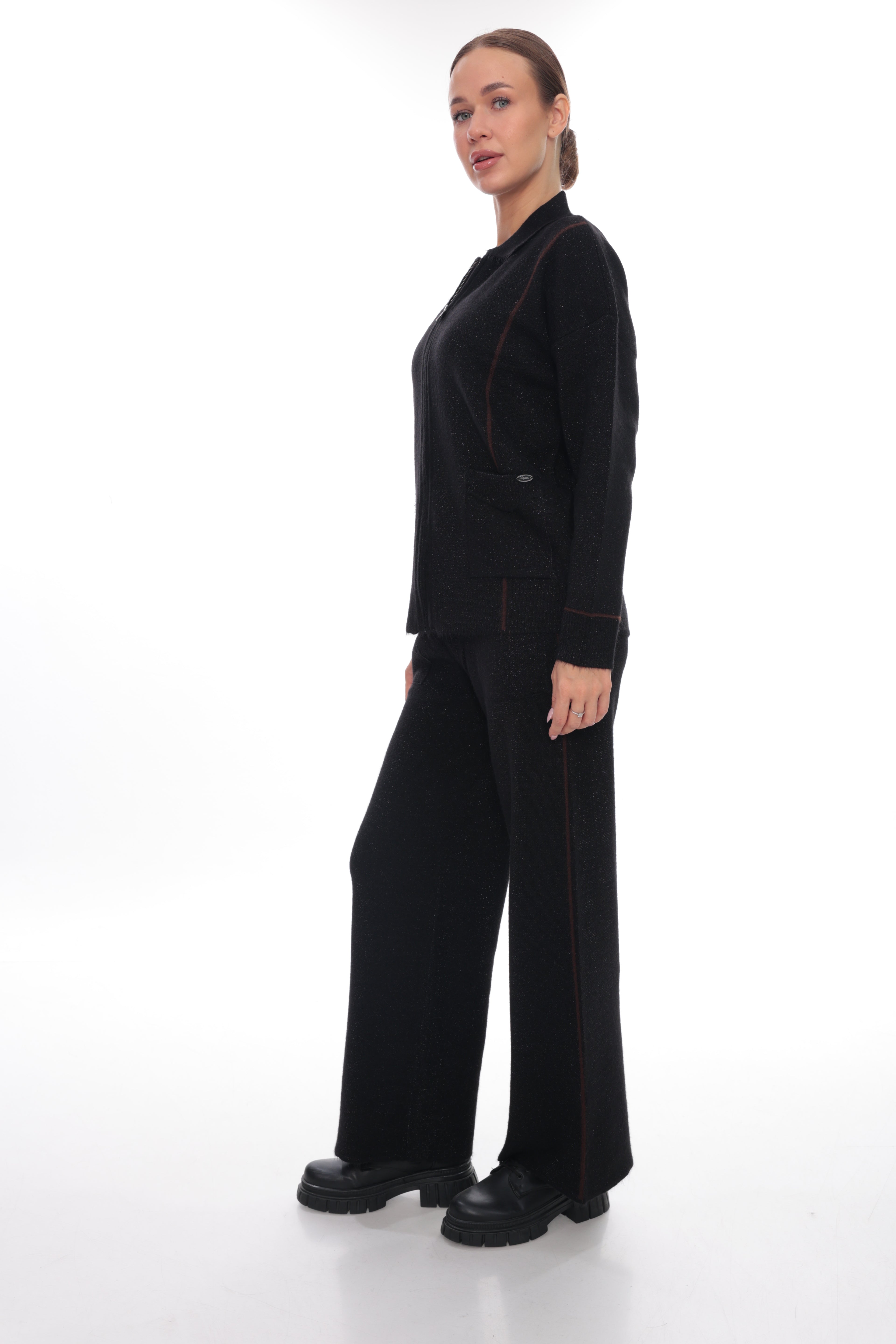 A woman modeling a black two-piece outfit consisting of a long-sleeve collared top and matching wide-leg trousers, both featuring subtle red contrast stitching, photographed against a seamless white studio background.