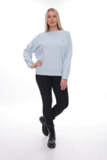 A woman modeling a light blue long-sleeve crewneck sweater with a small gold embroidered logo on the left chest, paired with black leggings and black boots, against a white studio background.