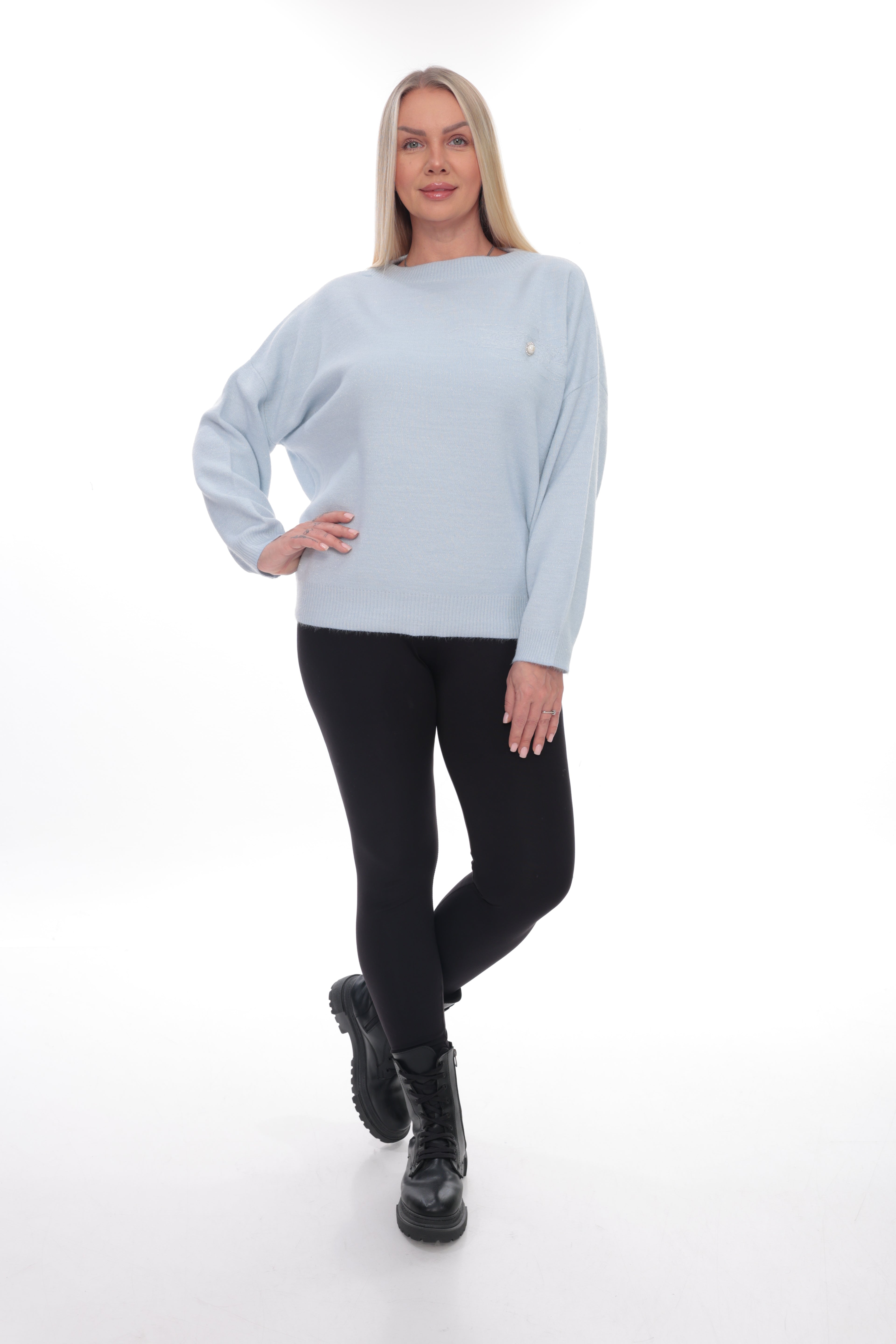 A woman modeling a light blue long-sleeve crewneck sweater with a small gold embroidered logo on the left chest, paired with black leggings and black boots, against a white studio background.