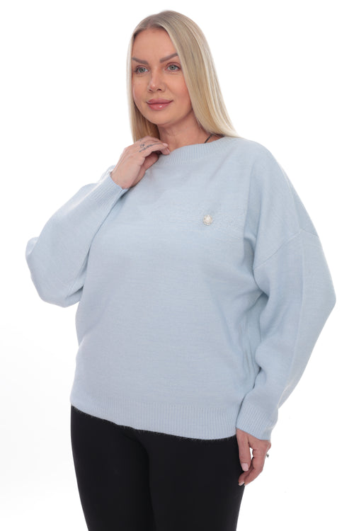 A woman modeling a light blue knit sweater with a small white embroidered logo on the left chest, paired with black pants, against a plain white background.