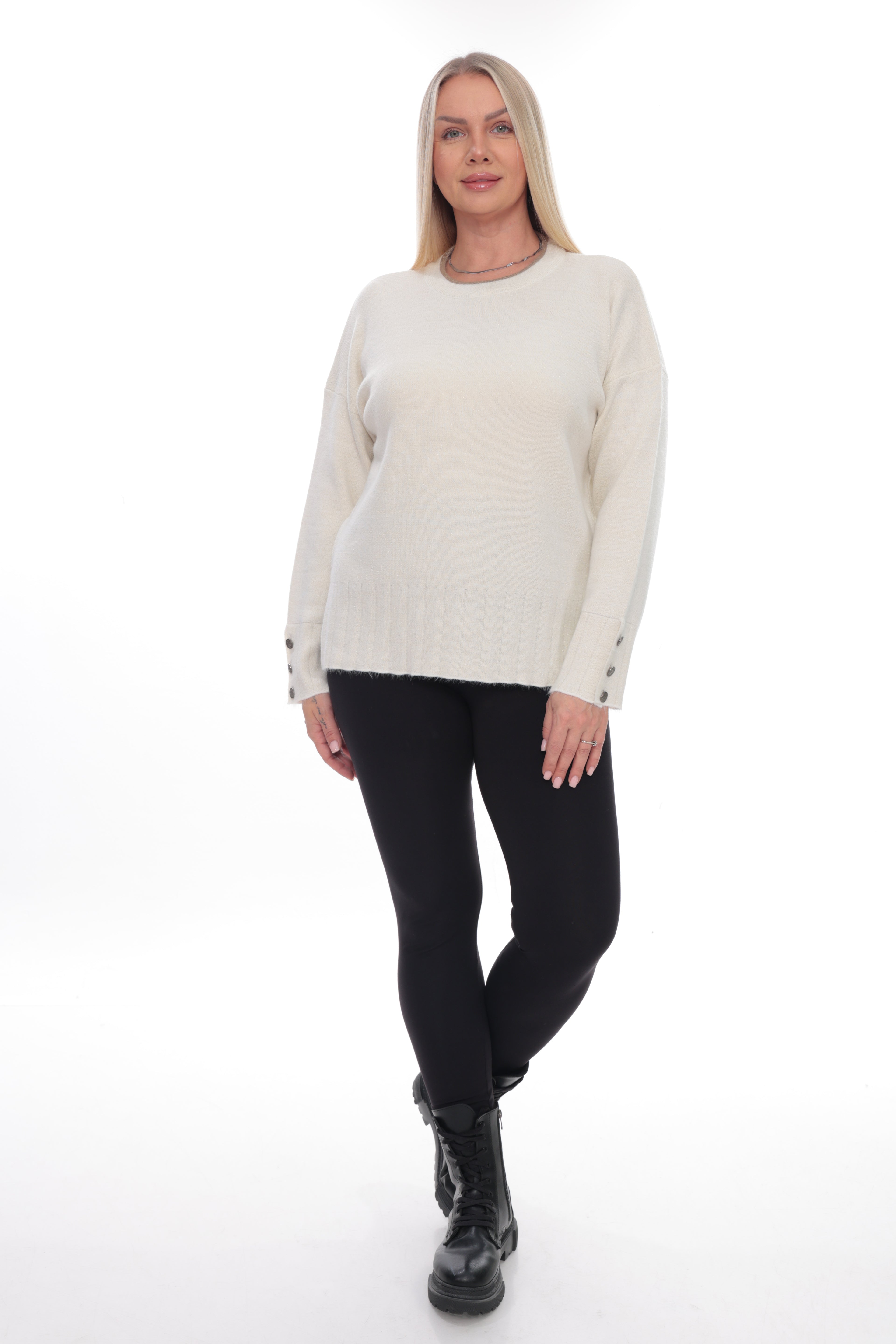 A woman modeling a cream-colored knit sweater with black pants and black boots, standing against a solid white studio background.