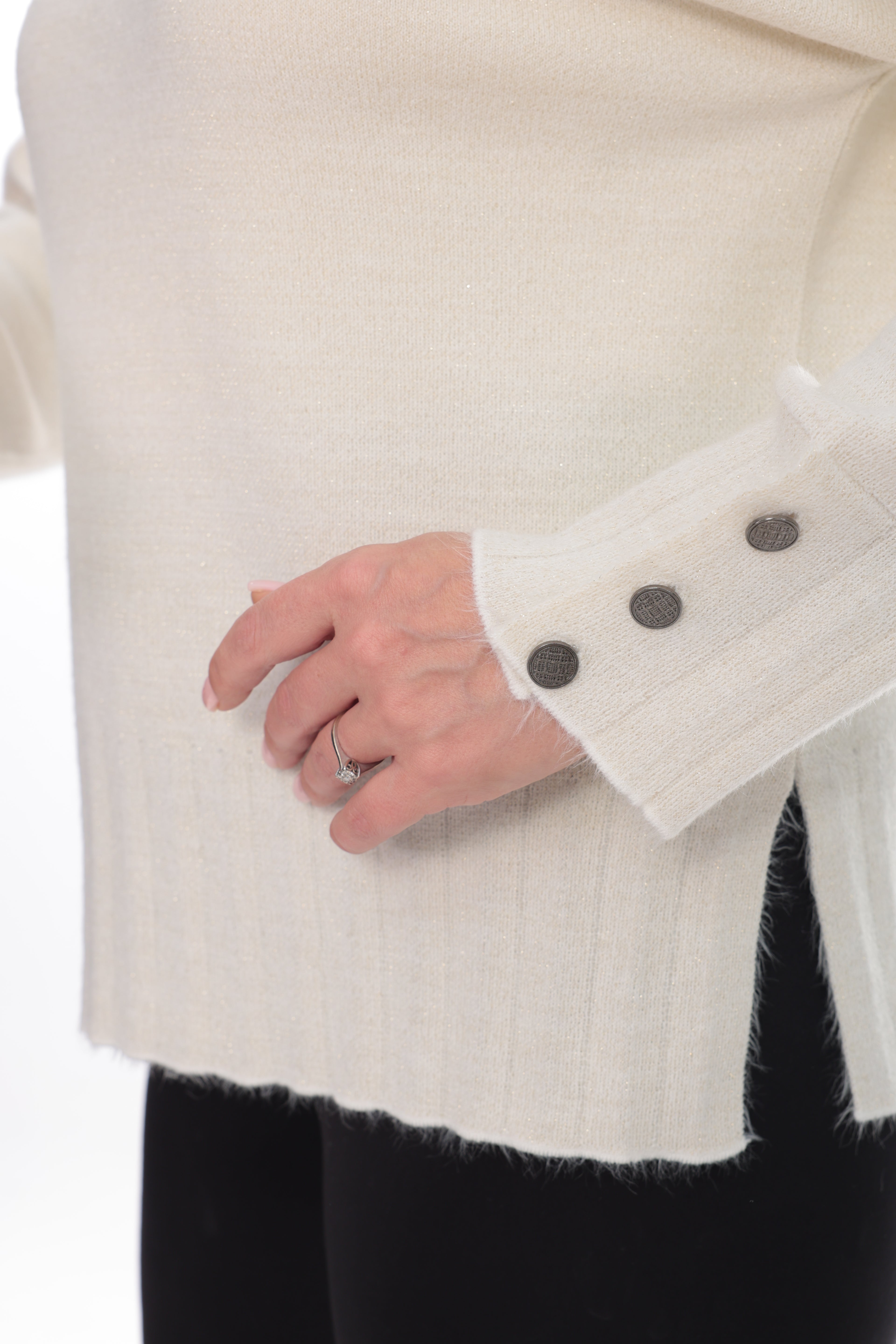 Close-up of a person wearing a cream ribbed knit sweater with a frayed hem and three dark buttons on the cuff, paired with black pants.