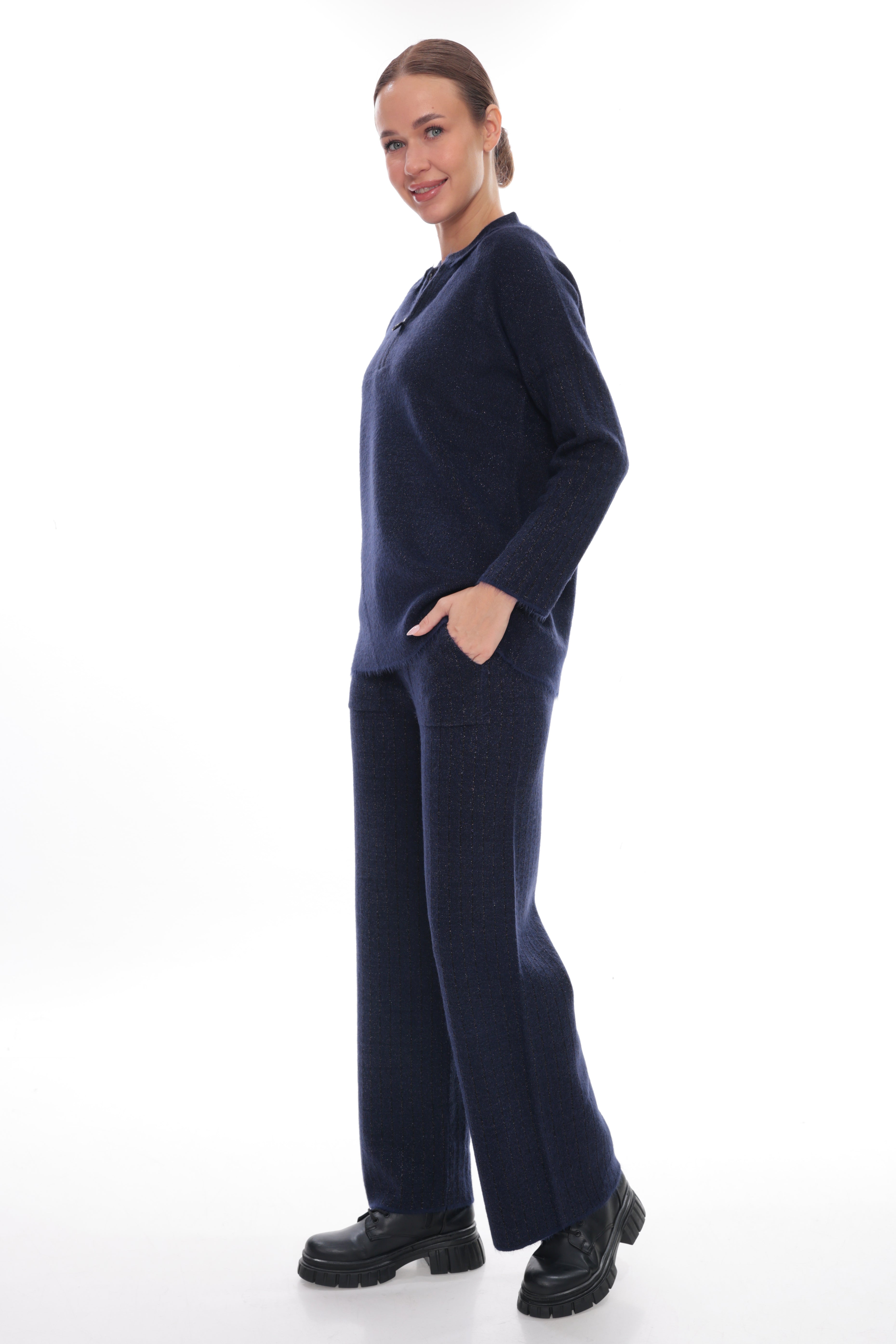 A woman modeling a matching navy blue knit lounge set consisting of a long-sleeve top and wide-leg trousers, paired with black platform shoes, against a solid white studio background.