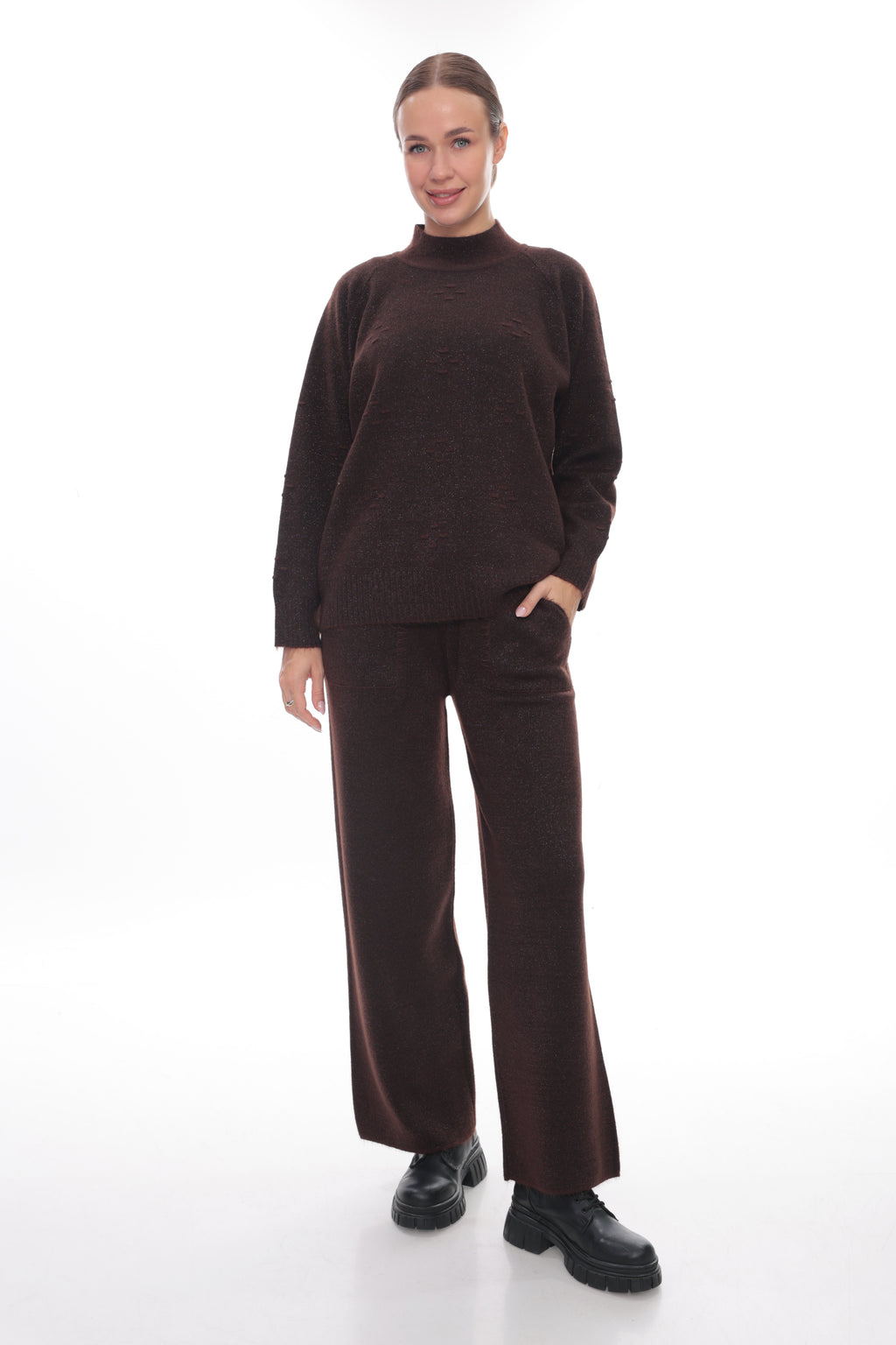 A woman modeling a dark brown knit two-piece set consisting of a mock neck long-sleeve sweater and matching wide-leg trousers, paired with black shoes, against a solid white studio background.