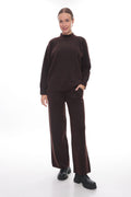 A woman modeling a dark brown knit two-piece set consisting of a mock neck long-sleeve sweater and matching wide-leg trousers, paired with black shoes, against a solid white studio background.