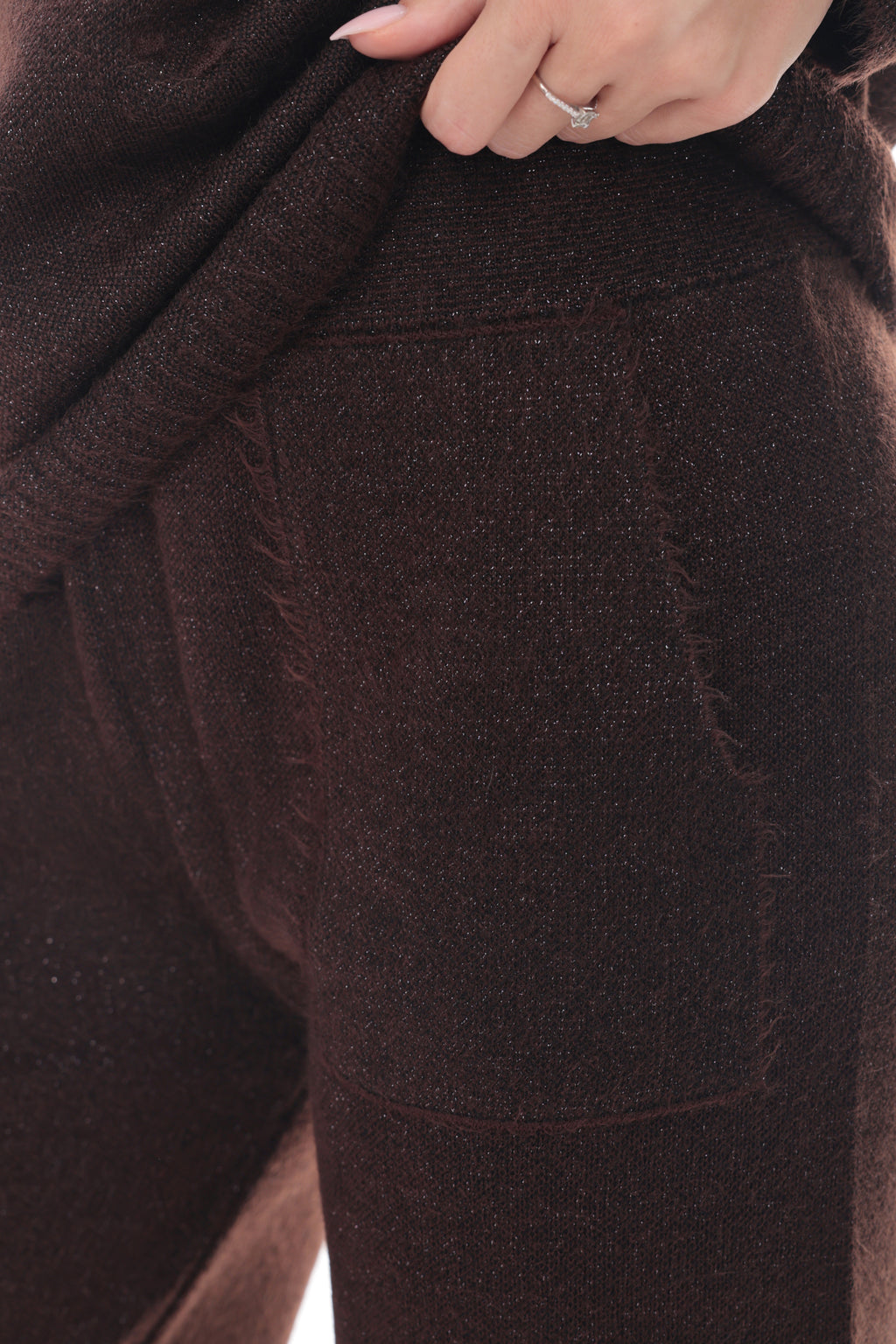 Close-up of a person wearing dark brown knit pants, pulling the fabric slightly to show texture and a side pocket, with a hand and ring visible at the top.