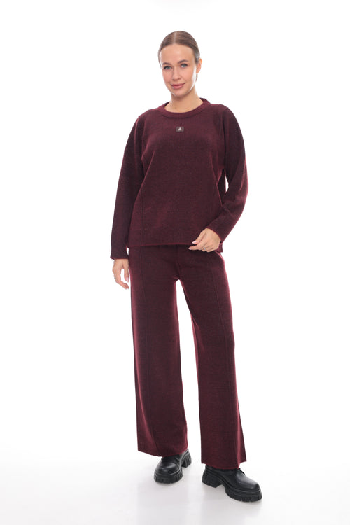 A woman modeling a matching burgundy corduroy lounge set, consisting of a long-sleeve crewneck top and wide-leg trousers, paired with black shoes, against a plain white studio background.