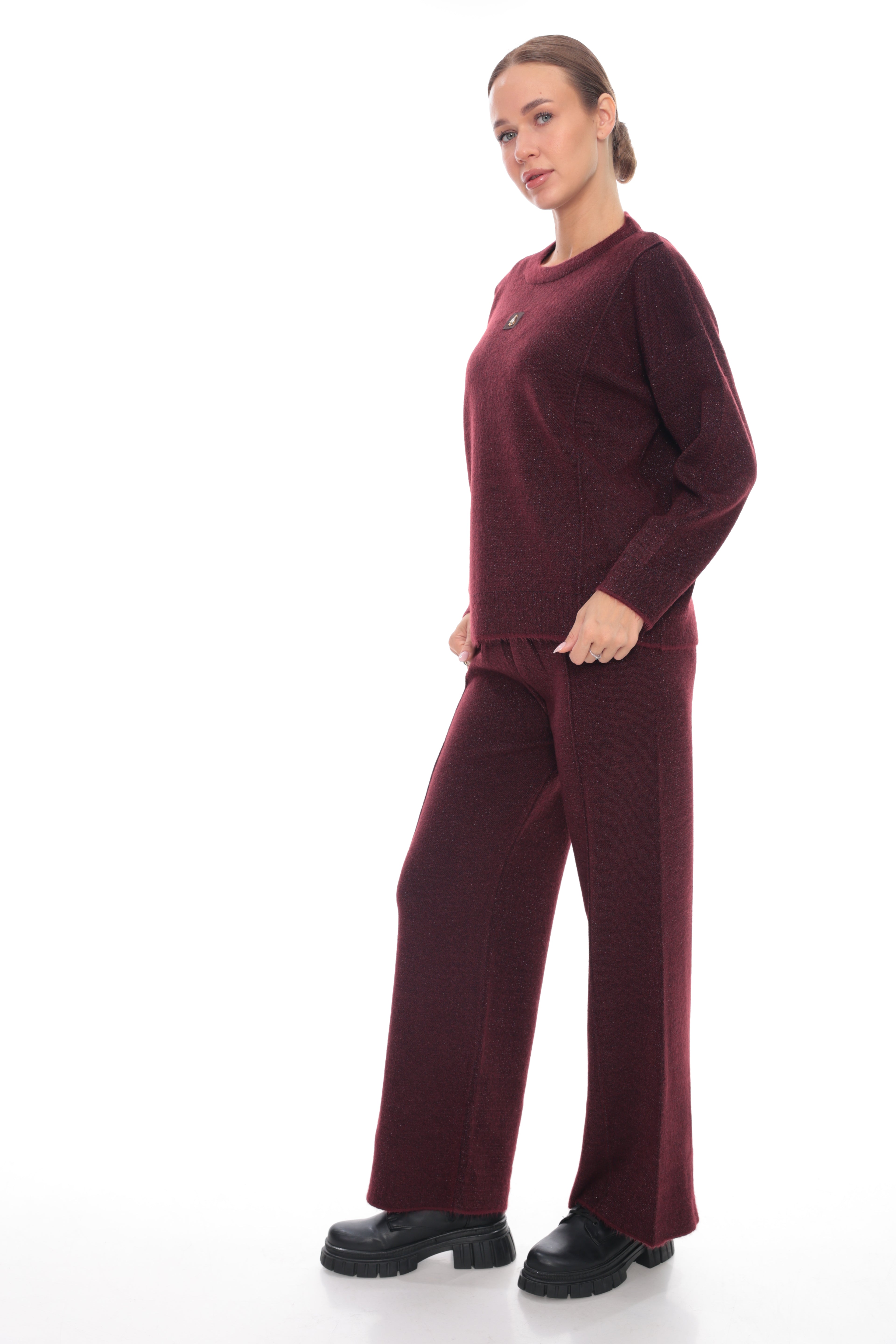 A woman modeling a matching burgundy knit loungewear set consisting of a long-sleeve top and wide-leg pants, paired with black boots against a plain white background.