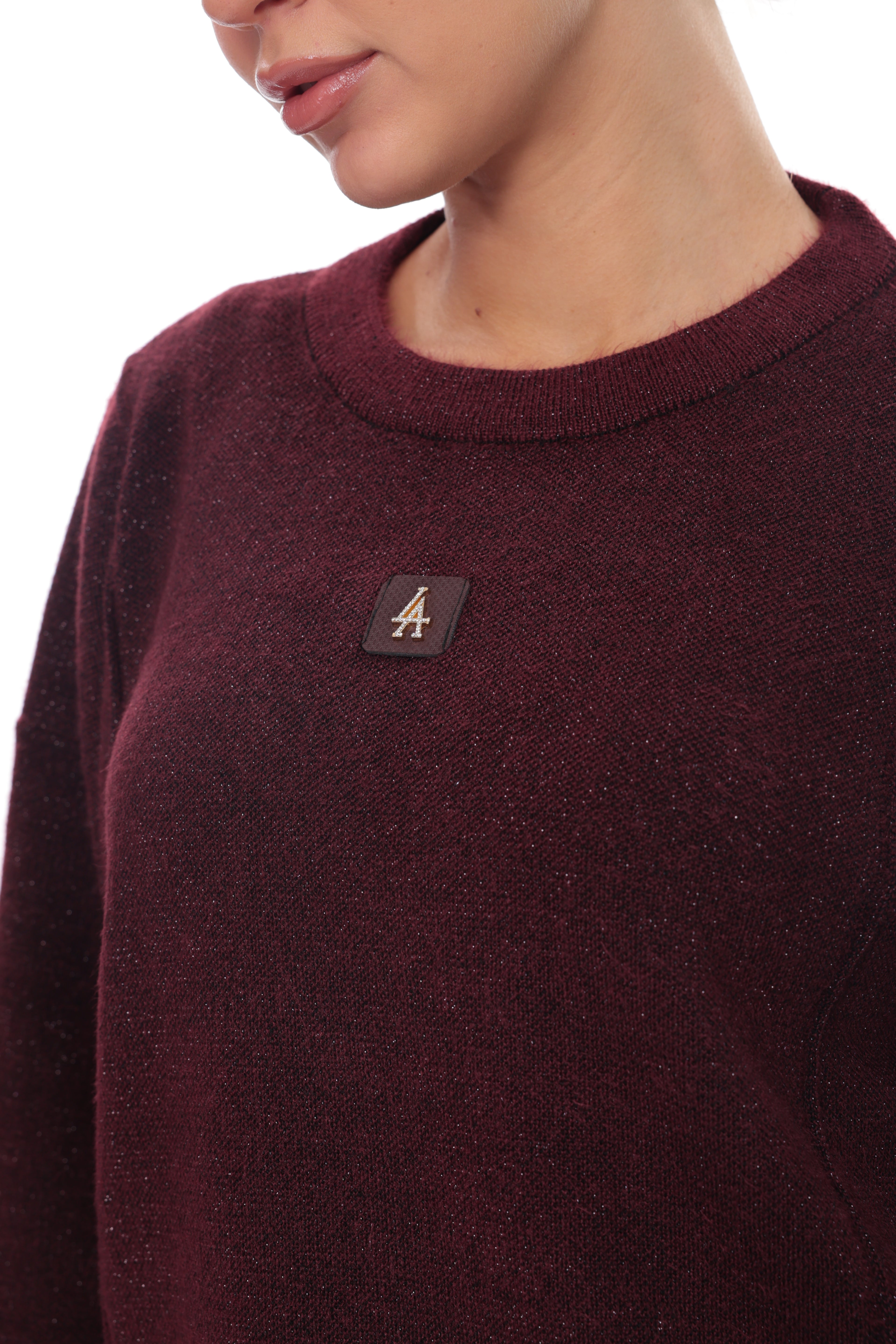 Close-up of a person wearing a dark burgundy knit crewneck sweater with a small black rectangular chest patch featuring a gold letter 'A'.