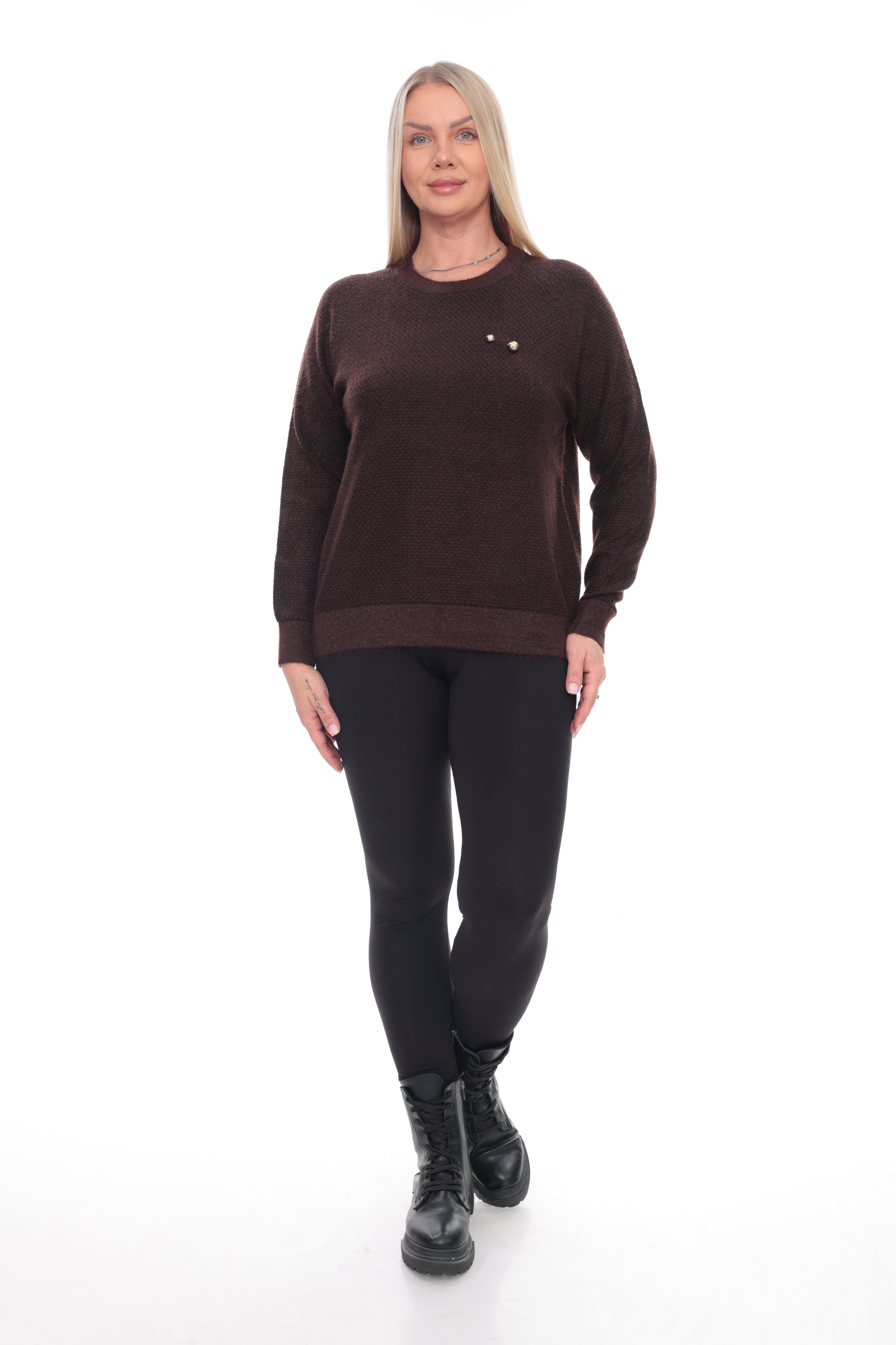 A woman modeling a dark brown crewneck sweater with a small gold chest emblem, paired with black leggings and black boots, against a plain white background.