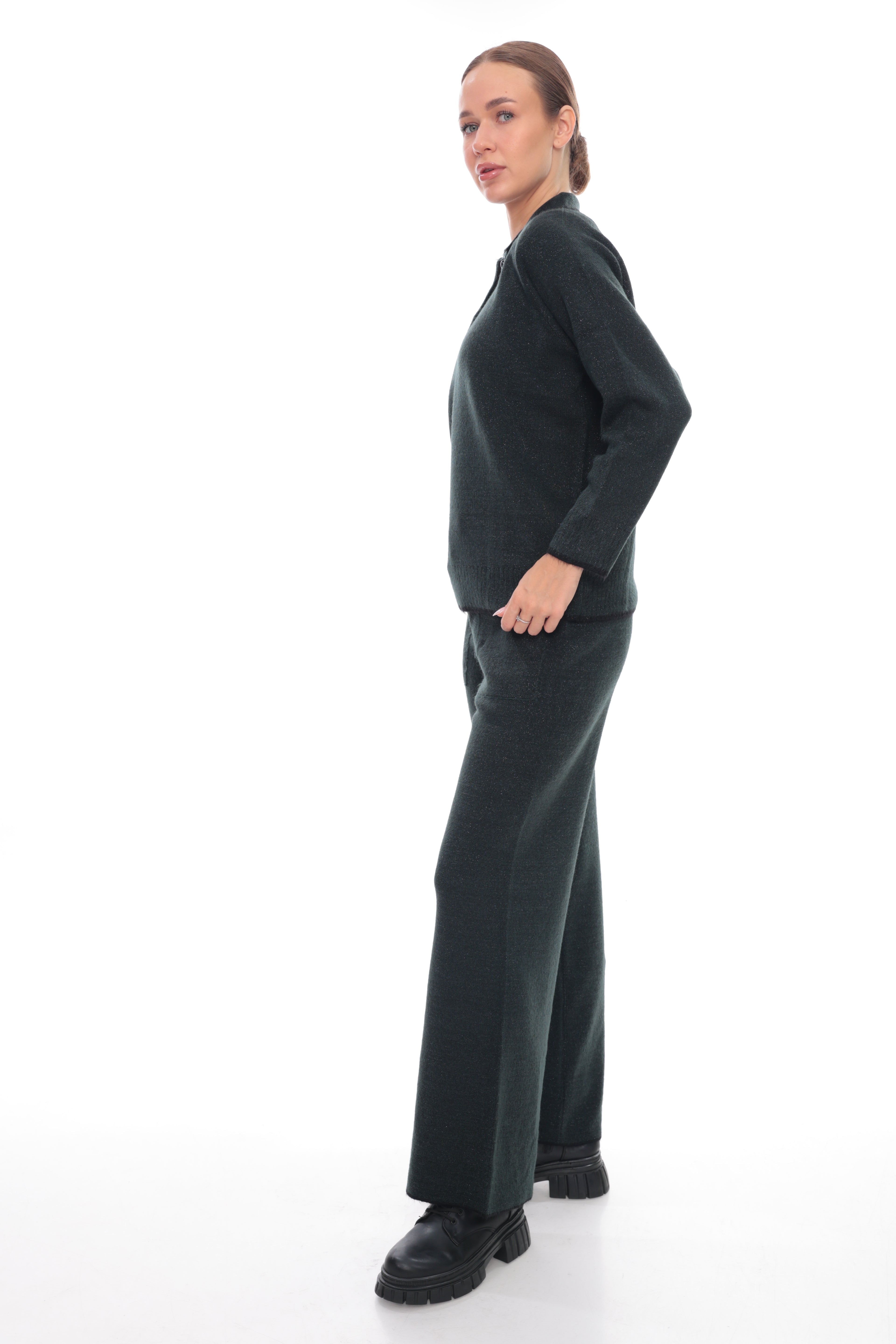 A woman modeling a dark green knit two-piece set consisting of a long-sleeve top and matching wide-leg trousers, paired with black shoes, against a solid white studio background.