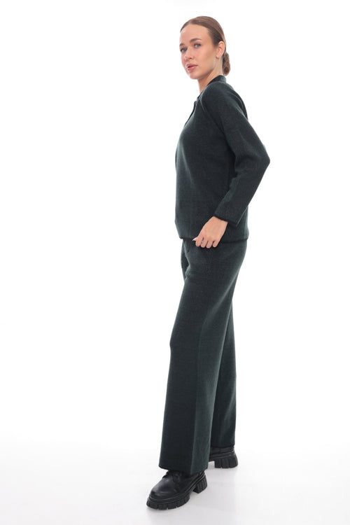 A woman modeling a dark green knit two-piece set consisting of a long-sleeve top and matching wide-leg trousers, paired with black shoes, against a solid white studio background.
