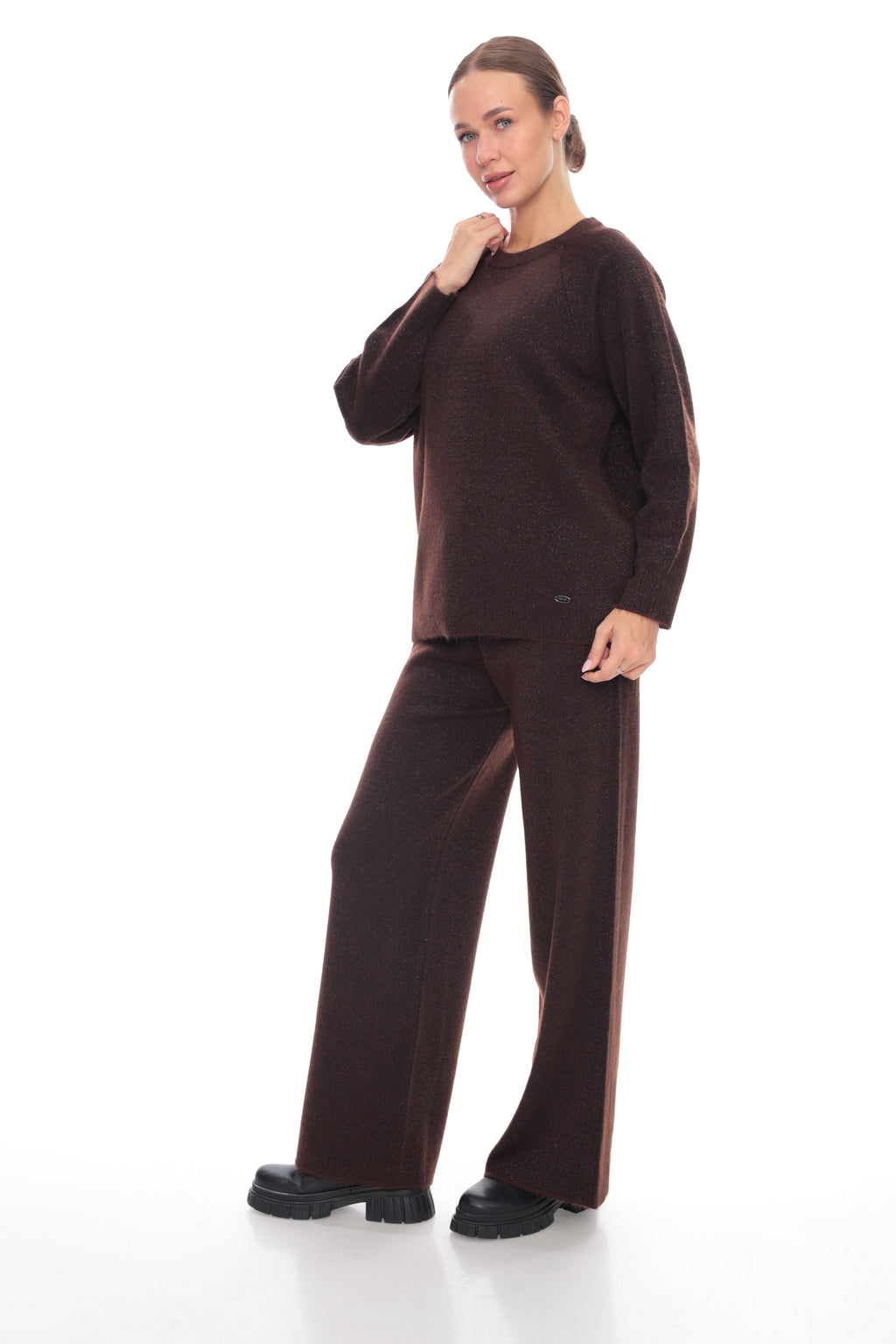 A woman modeling a matching dark brown knit lounge set consisting of a long-sleeve crewneck sweater and wide-leg trousers, paired with black platform shoes against a white studio background.