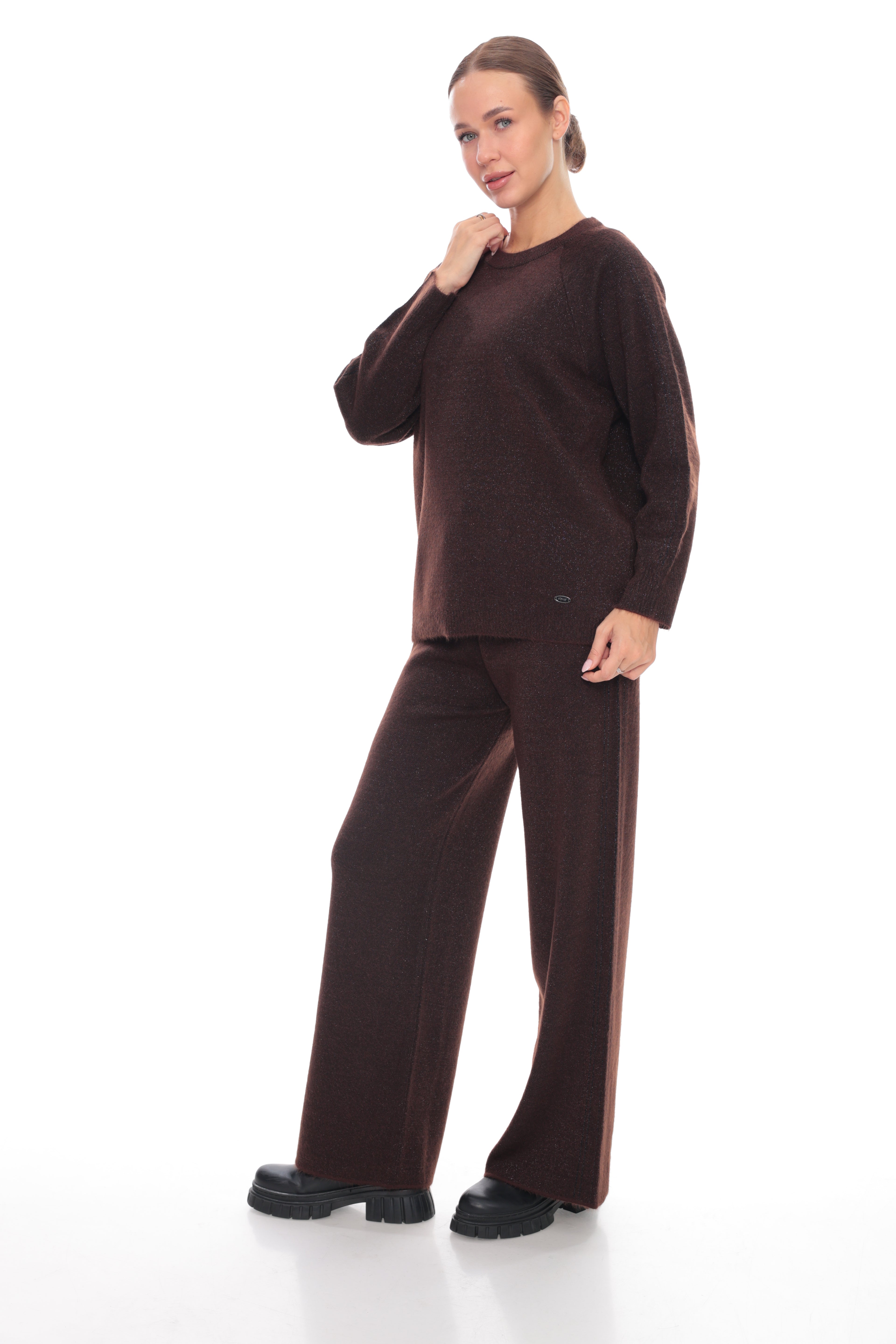 A woman modeling a matching dark brown knit lounge set consisting of a long-sleeve crewneck sweater and wide-leg trousers, paired with black platform shoes against a white studio background.