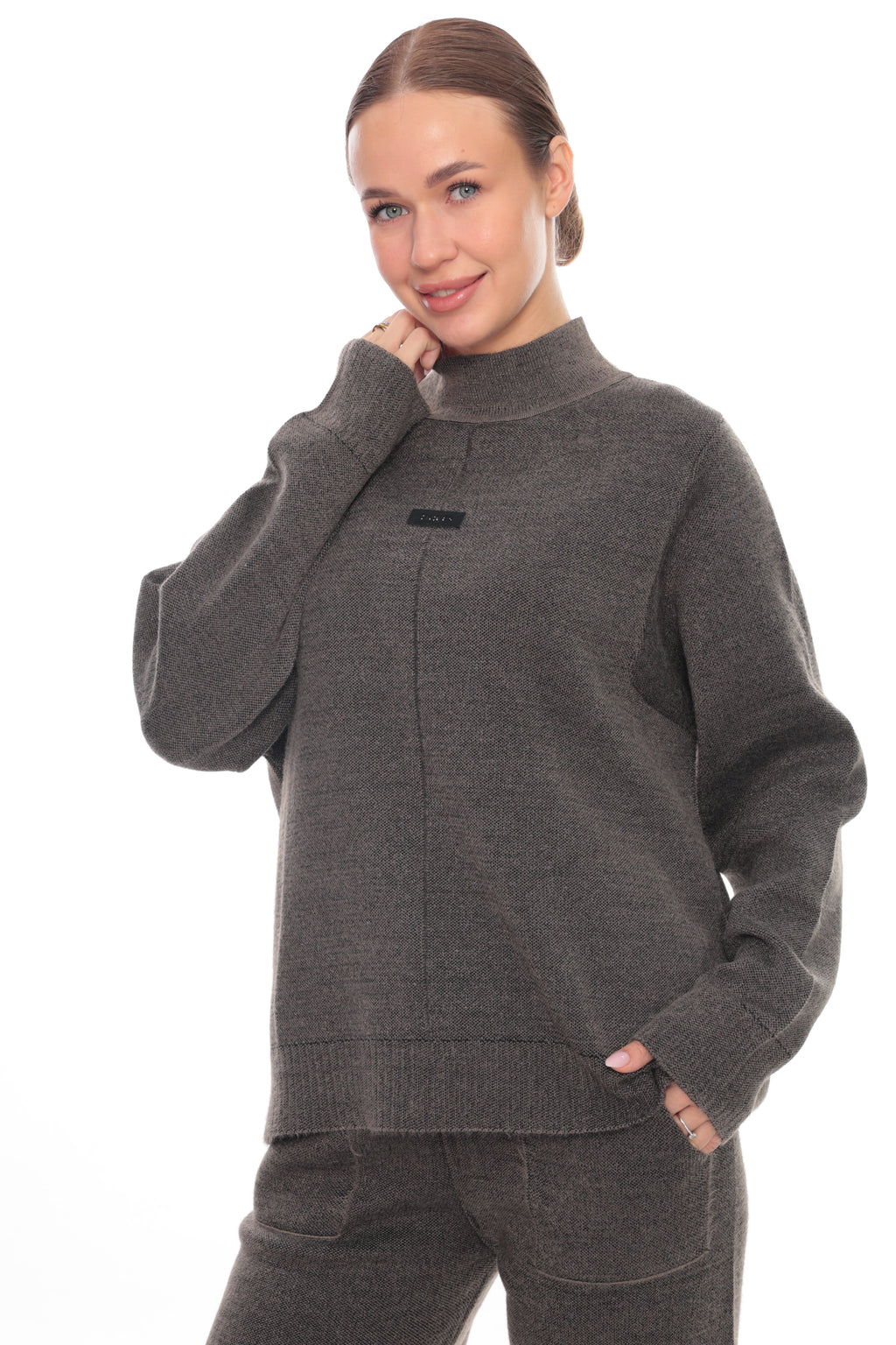A woman wearing a dark gray ribbed knit mock-neck sweater with a small black brand label on the chest, paired with matching gray pants, against a plain white background.