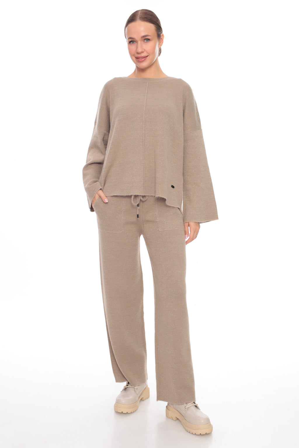 A woman modeling a matching beige knit lounge set consisting of a cropped long-sleeve top and wide-leg pants, paired with beige sneakers, against a plain white studio background.