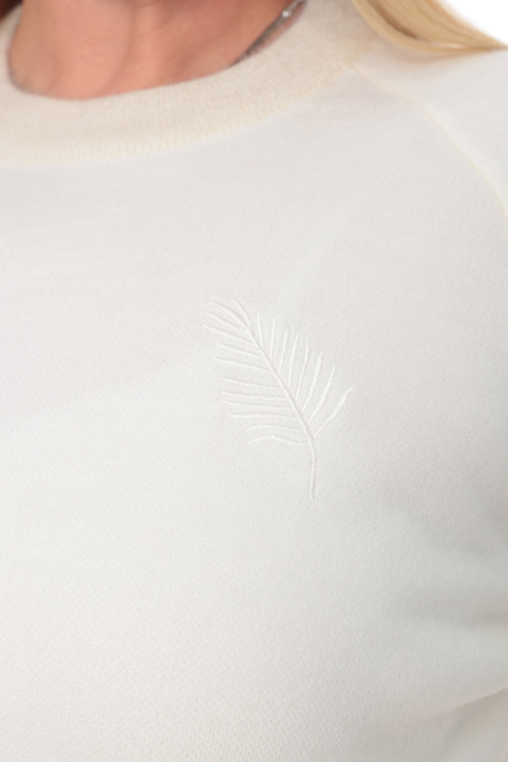 Close-up of a cream-colored crewneck sweater featuring a subtle, tonal embroidered leaf motif on the left chest, worn by a model.