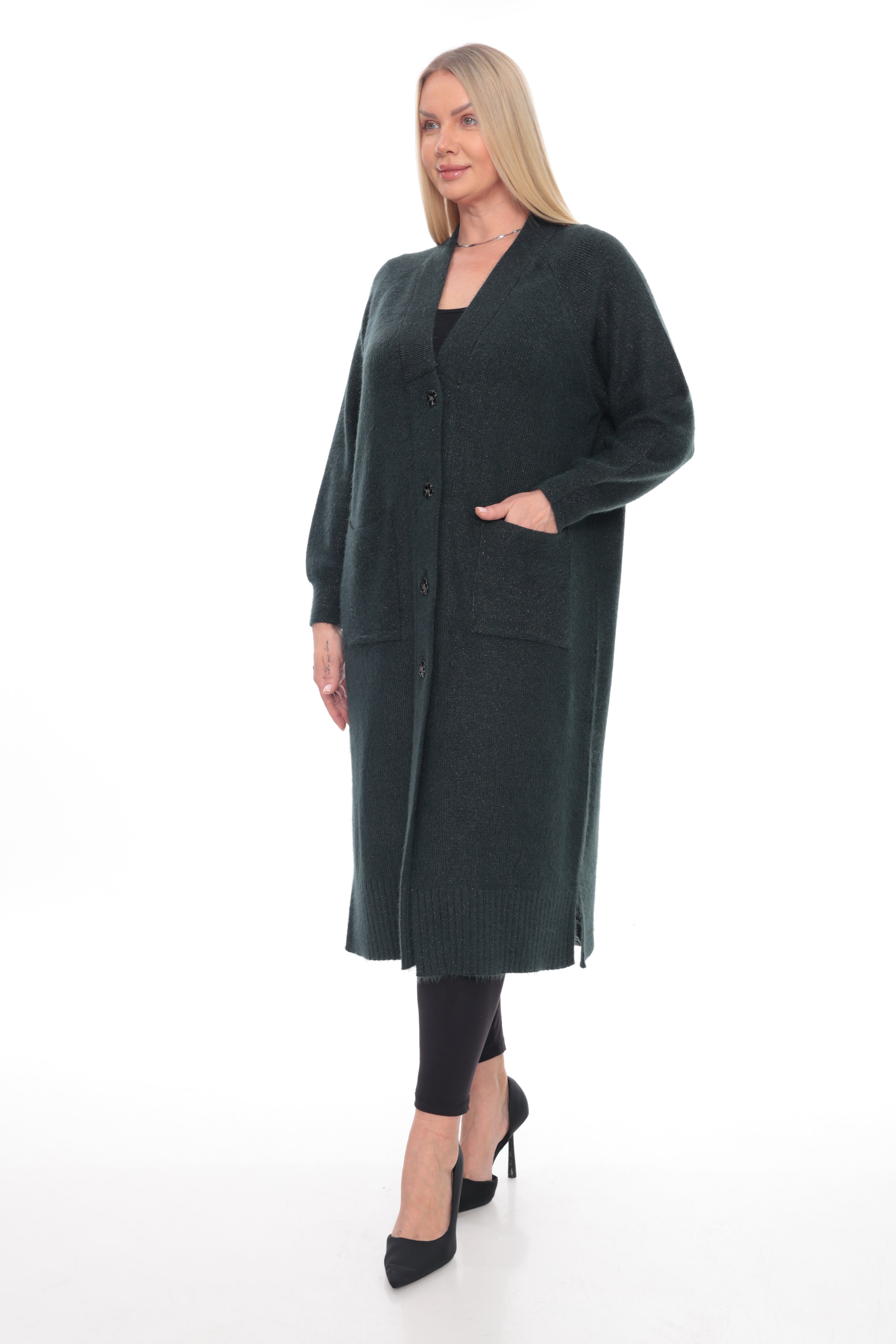 A woman modeling a long, dark green knit cardigan with front pockets and buttons, paired with black leggings and black high heels, against a plain white studio background.