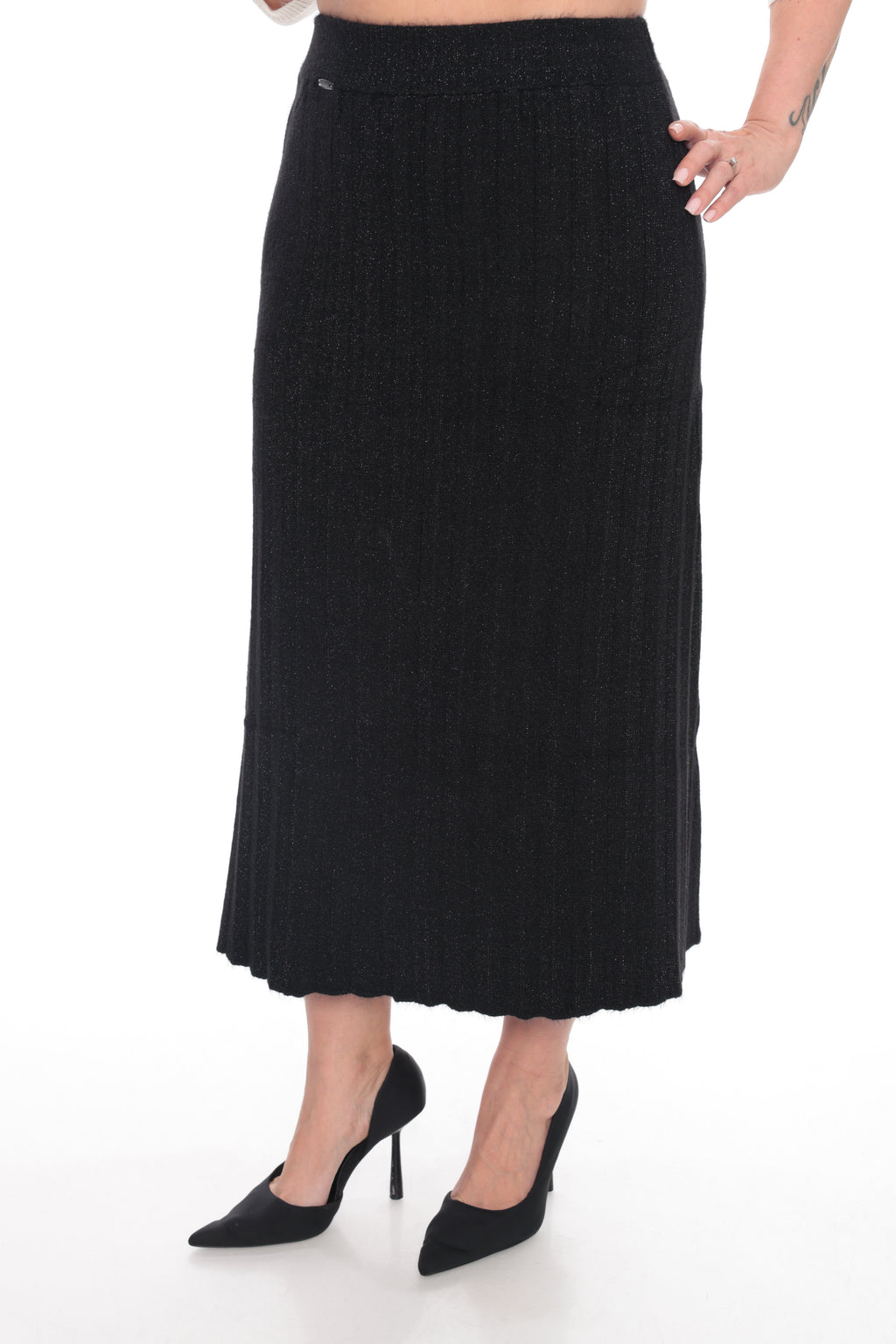 A black ribbed knit midi skirt worn by a model, paired with black pointed-toe heels, shown against a plain white studio background.