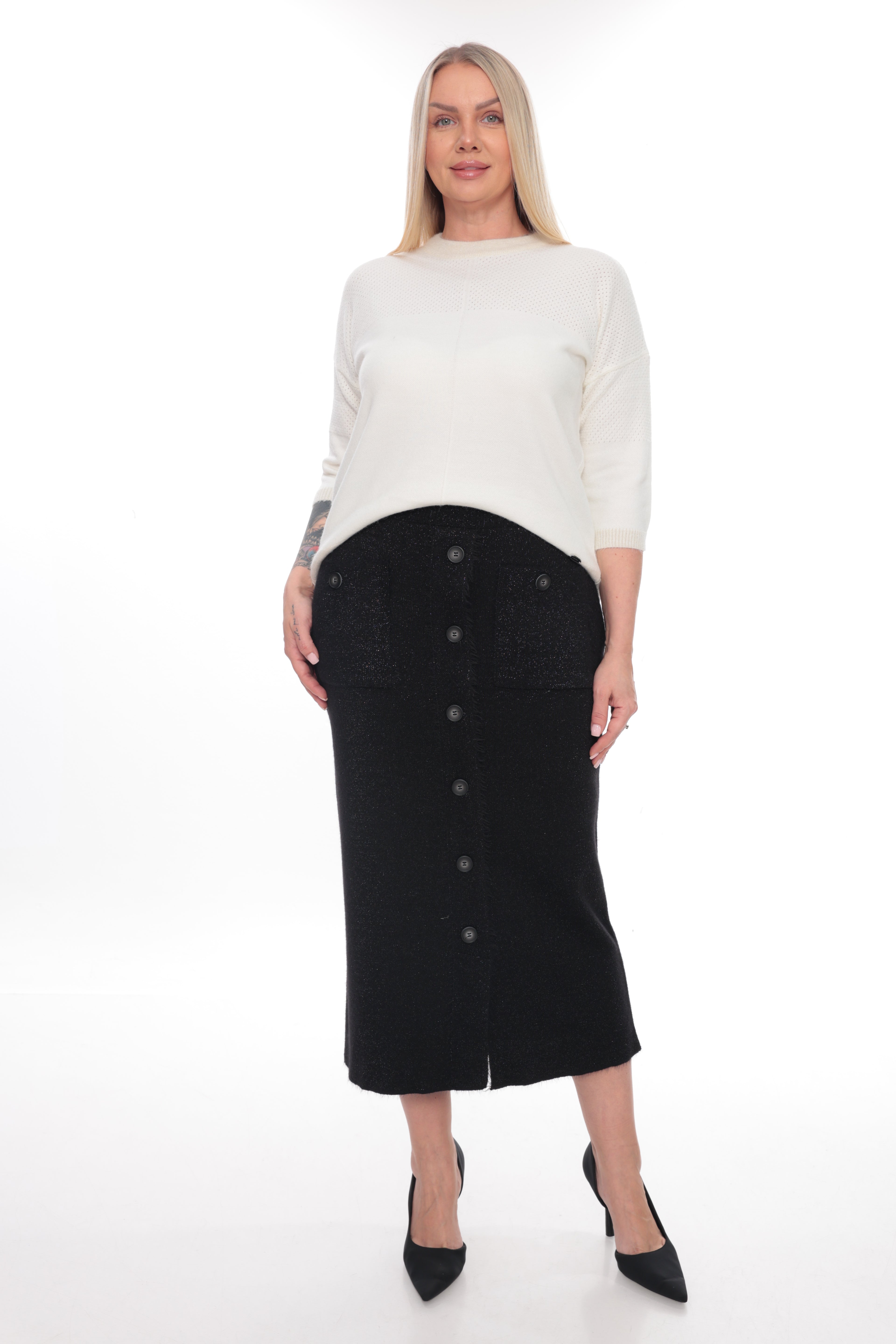 A woman modeling a black midi skirt with front buttons and textured pocket details, paired with a white long-sleeve top and black pointed heels, against a white studio background.