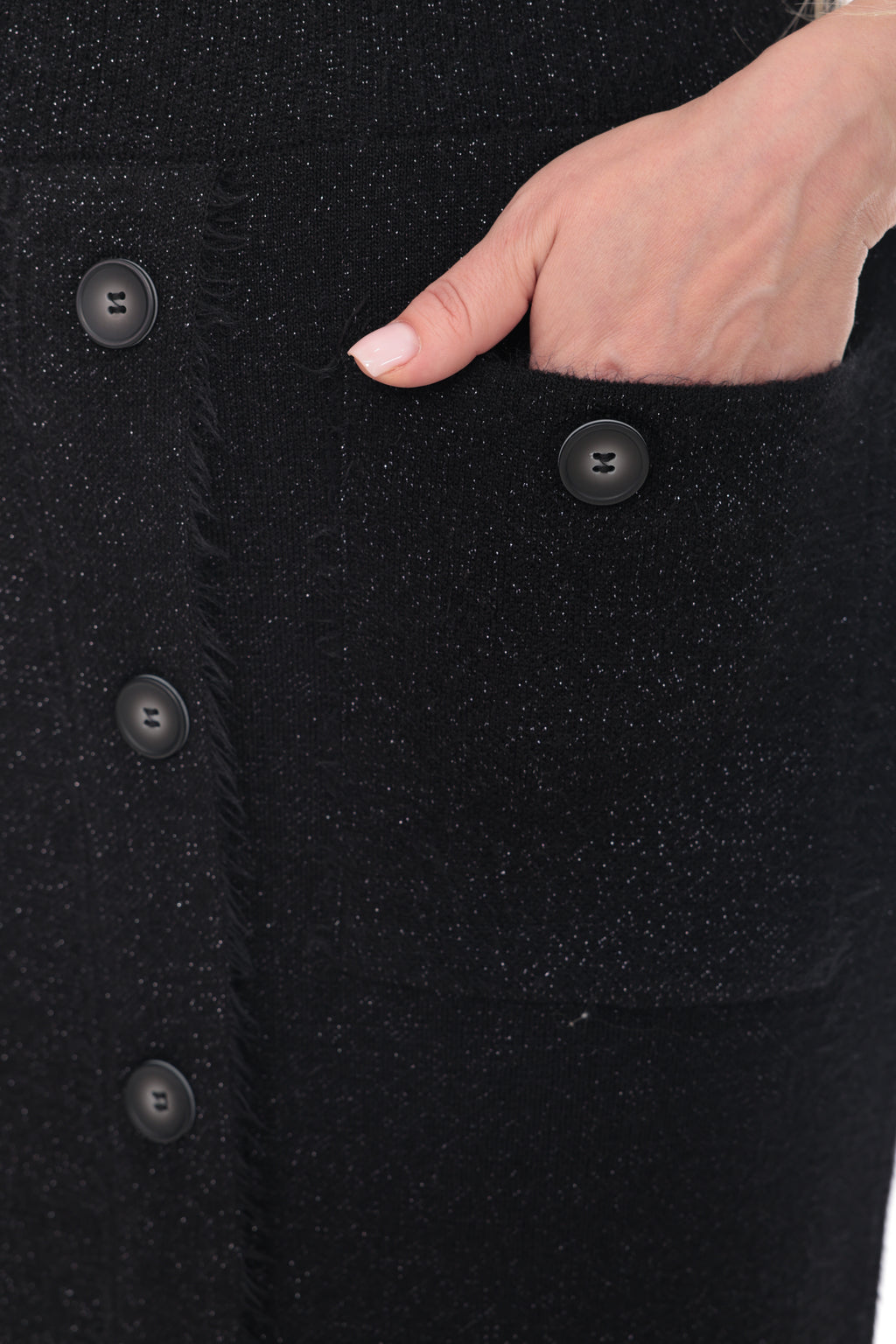 Close-up of a black textured skirt with a hand reaching into a front pocket, showing dark buttons and a fringed edge.