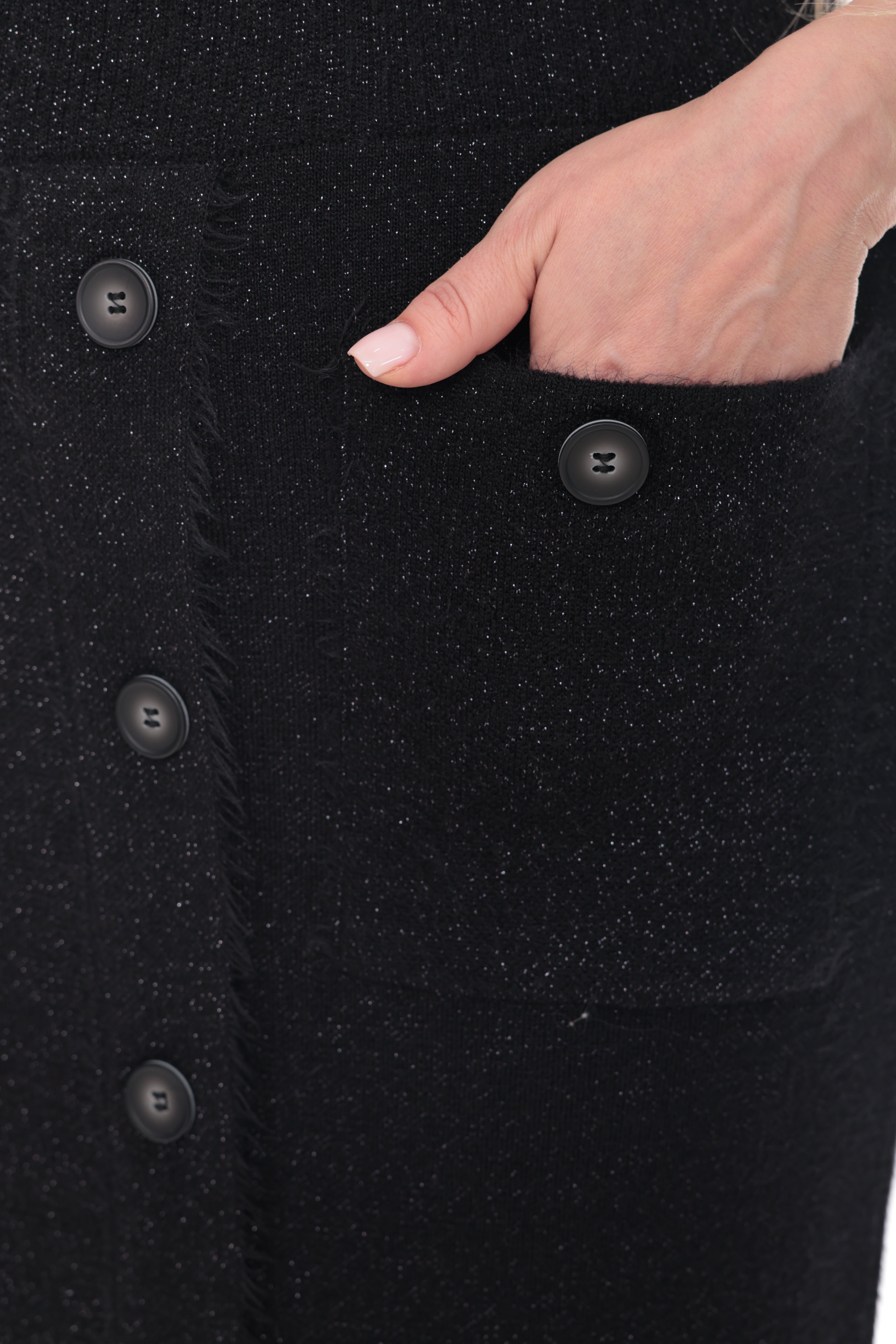 Close-up of a black textured skirt with a hand reaching into a front pocket, showing dark buttons and a fringed edge.